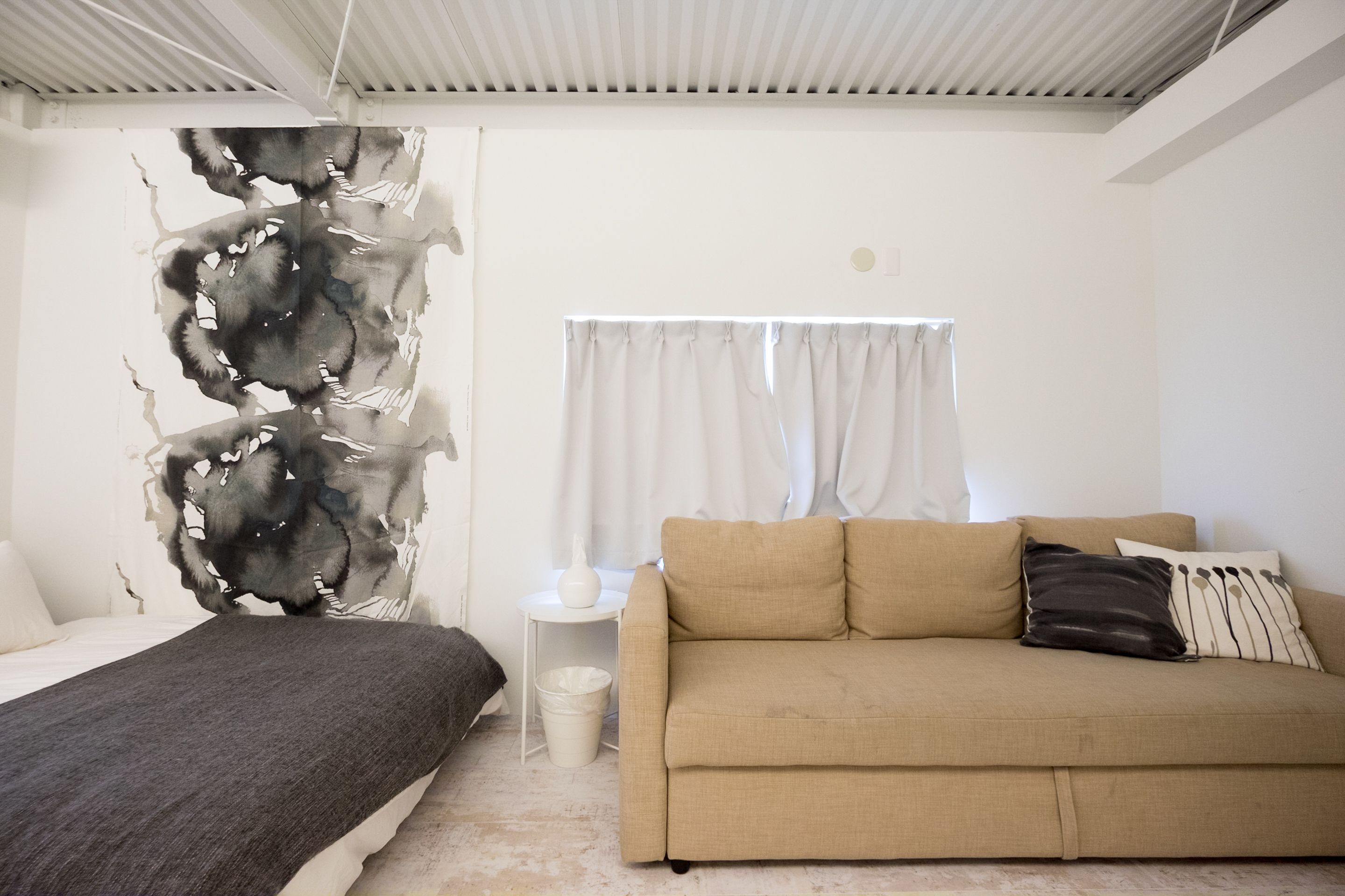A bedroom with a bed, a sofa, a wall art piece, a window with curtains, and a small table with a vase.