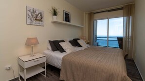 Une chambre d’hôtel avec un lit, une table de chevet, une lampe et une vue sur la mer, visible à travers une grande fenêtre.