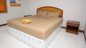 A neatly made bed with a beige bedspread and a rolled towel on top, a wooden headboard, a nightstand with a lamp, and a poster on the wall.
