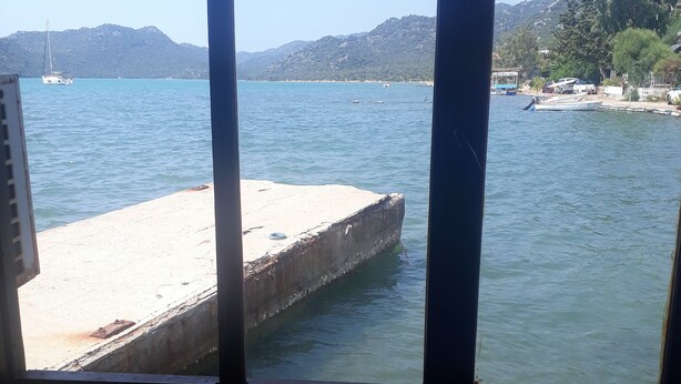 A view of a calm body of water with a concrete dock extending into it, surrounded by hills and a clear sky. - Kekova Restaurant & Pansiyon (Demre)