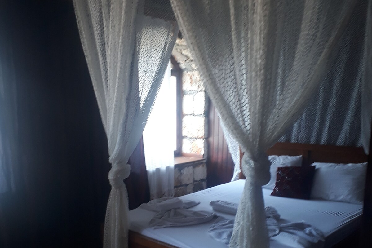 A bed with a wooden frame and white bedding, a window with a view of the outside, and a tiled floor.