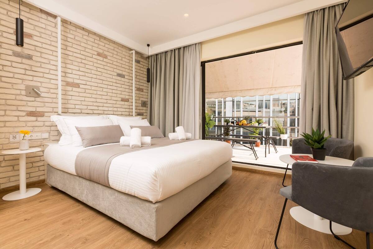 Quarto de hotel moderno com uma cama grande, uma mesa pequena, uma cadeira e uma varanda com vista para a cidade.