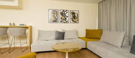 A modern living room with a sofa, a round glass coffee table, and two armchairs. There are three abstract paintings on the wall.