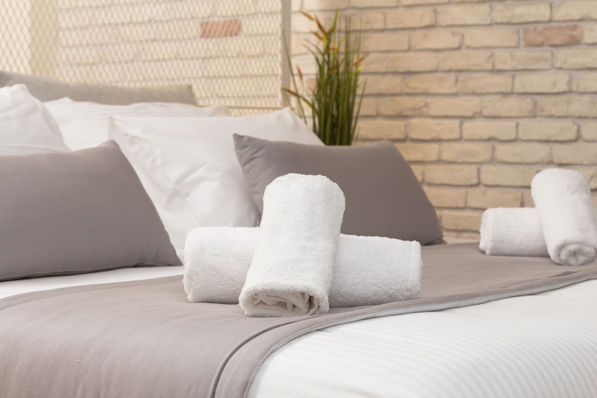 Cama bem arrumada com toalhas brancas dobradas, roupa de cama cinza e parede de tijolos ao fundo.