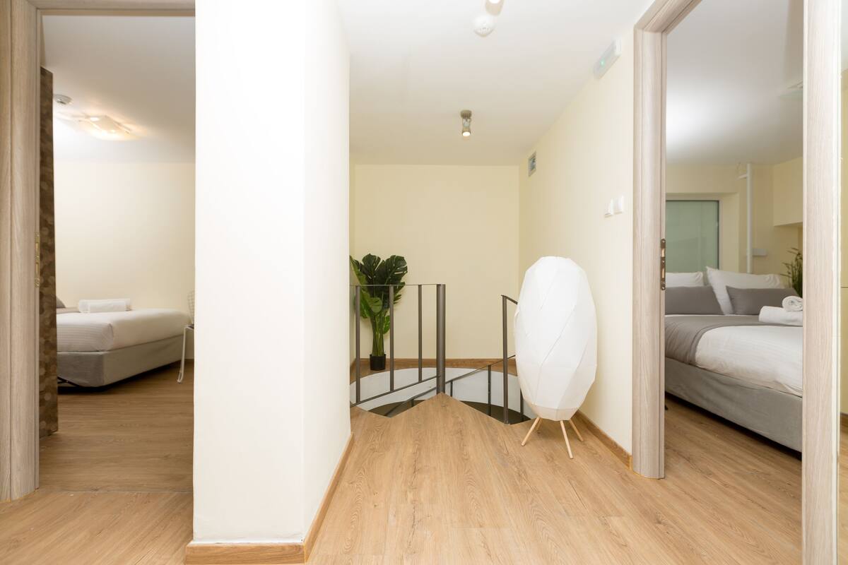 Quarto de hotel com chão de madeira, cama com roupa de cama cinza, uma cadeira branca e uma planta em vaso.