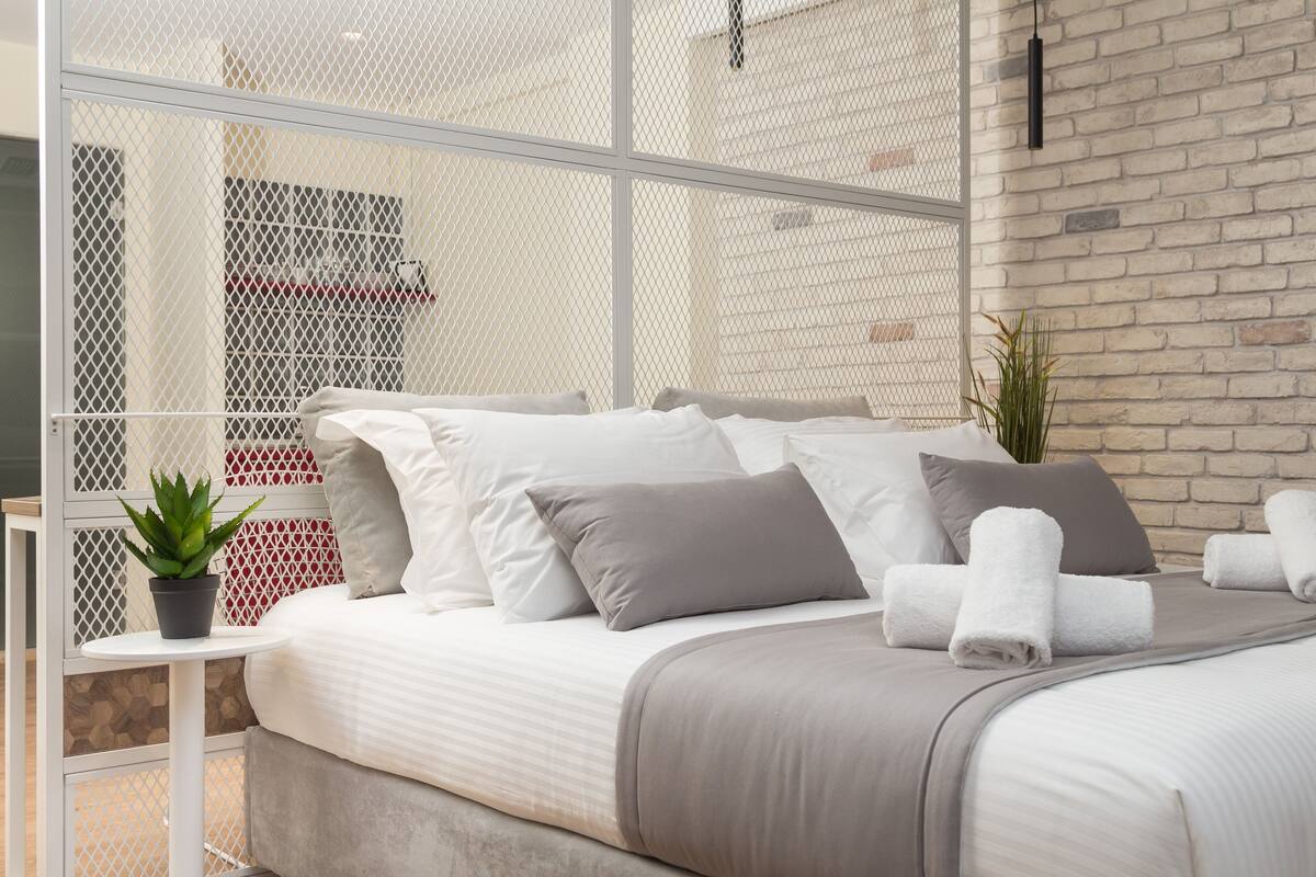 Quarto de hotel moderno com uma cama grande, uma mesinha com uma planta e vista através de uma janela com tela.