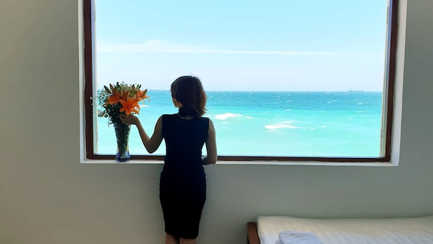 A room with a bed, a window with a view of the ocean, and a person holding a flower. - Mui Dinh Hotel (Ca Na)