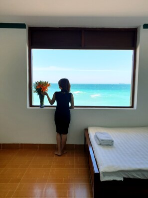 Una habitación con una cama, una ventana que da al océano y una persona sosteniendo una flor.