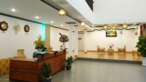 A hotel reception area with a wooden reception desk, potted plants, wall clocks, and a television screen. - Mui Dinh Hotel (Ca Na)