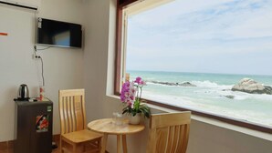 A room with a view of the ocean, a television mounted on the wall, a refrigerator, a table with chairs, and a potted plant. - Mui Dinh Hotel (Ca Na)