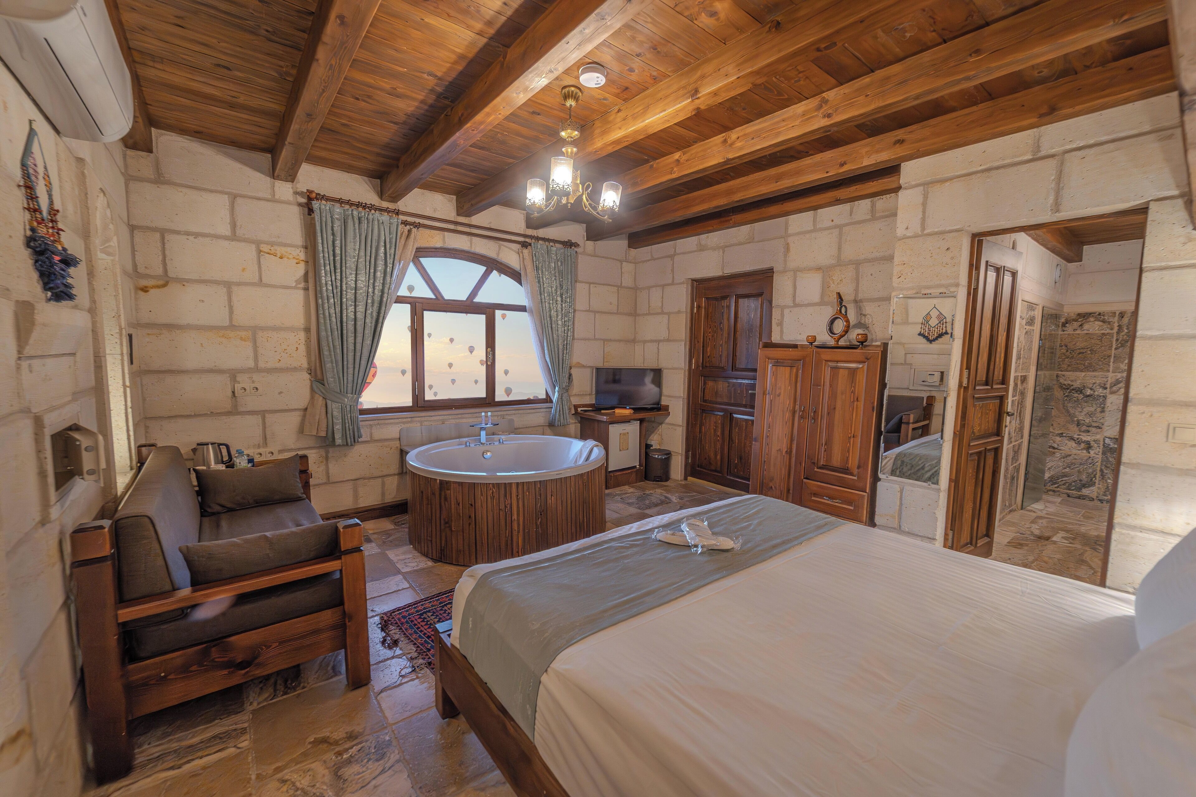 Un dormitorio acogedor con techo de madera, una cama grande, un área de descanso con sofá, una bañera redonda y una ventana con cortinas.