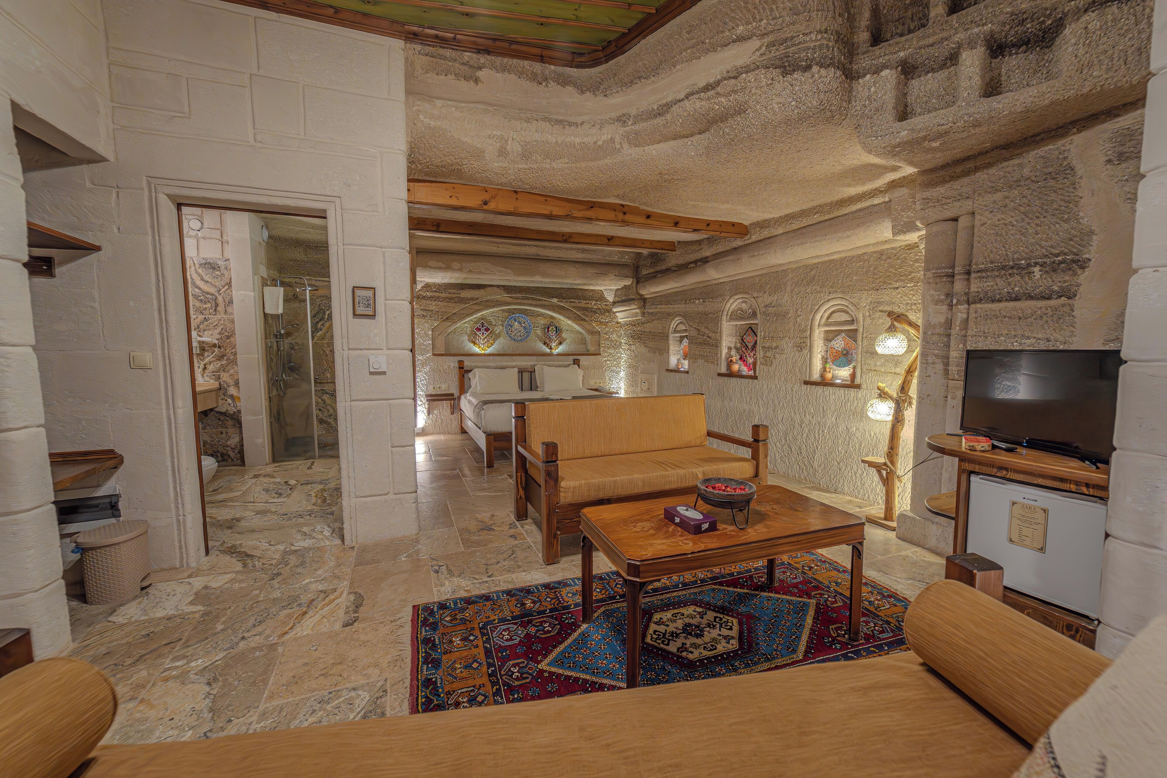 A spacious living area with a wooden sofa, a coffee table, and a television. The room features stone walls and a tiled floor.