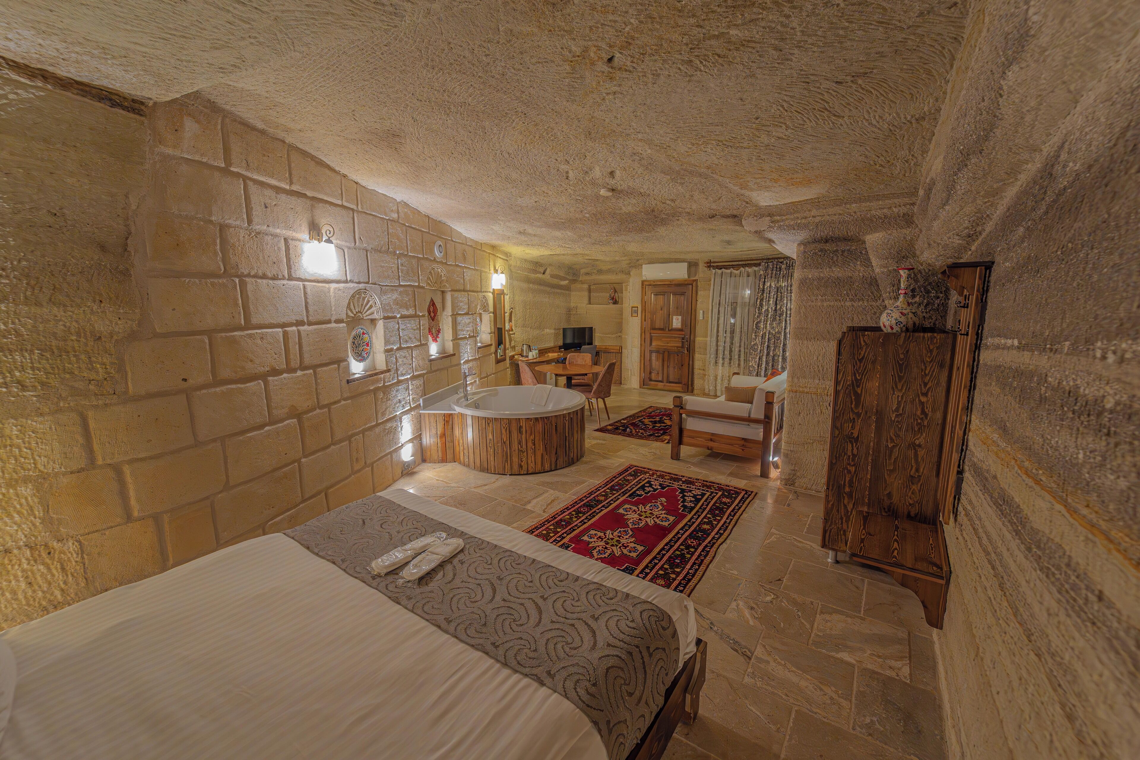 A stone-walled room with a bed, a round bathtub, a wooden cabinet, and a small table with chairs.