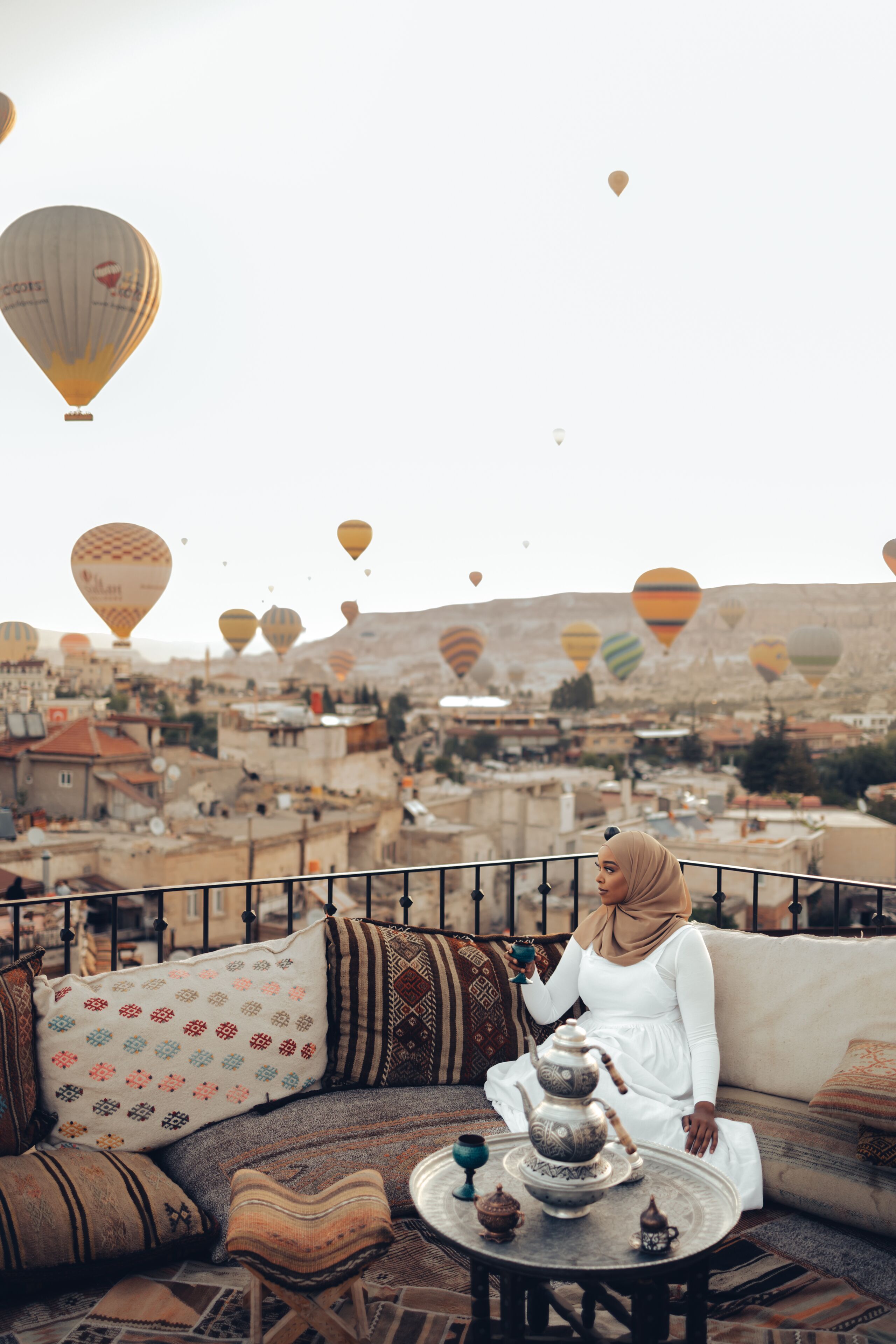 Eine Frau sitzt auf einem Balkon mit Blick auf eine Stadtlandschaft und dahinter schwebende Heißluftballons.