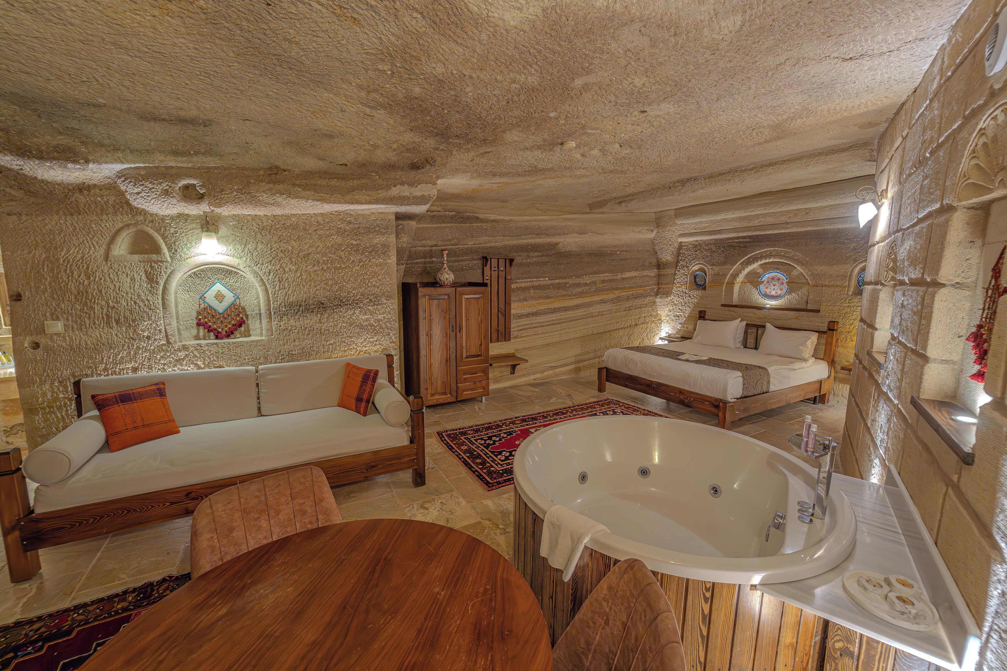A cozy, stone-walled room with a wooden sofa, a round wooden table, a large bathtub, and a bed with a wooden headboard.
