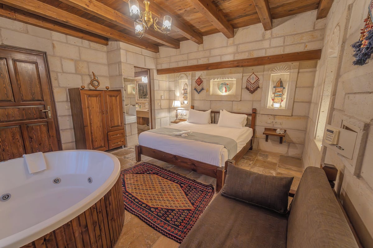 Une chambre à coucher rustique avec un grand lit, une tête de lit en bois, un coin salon avec un banc, une baignoire et un mur en pierre.