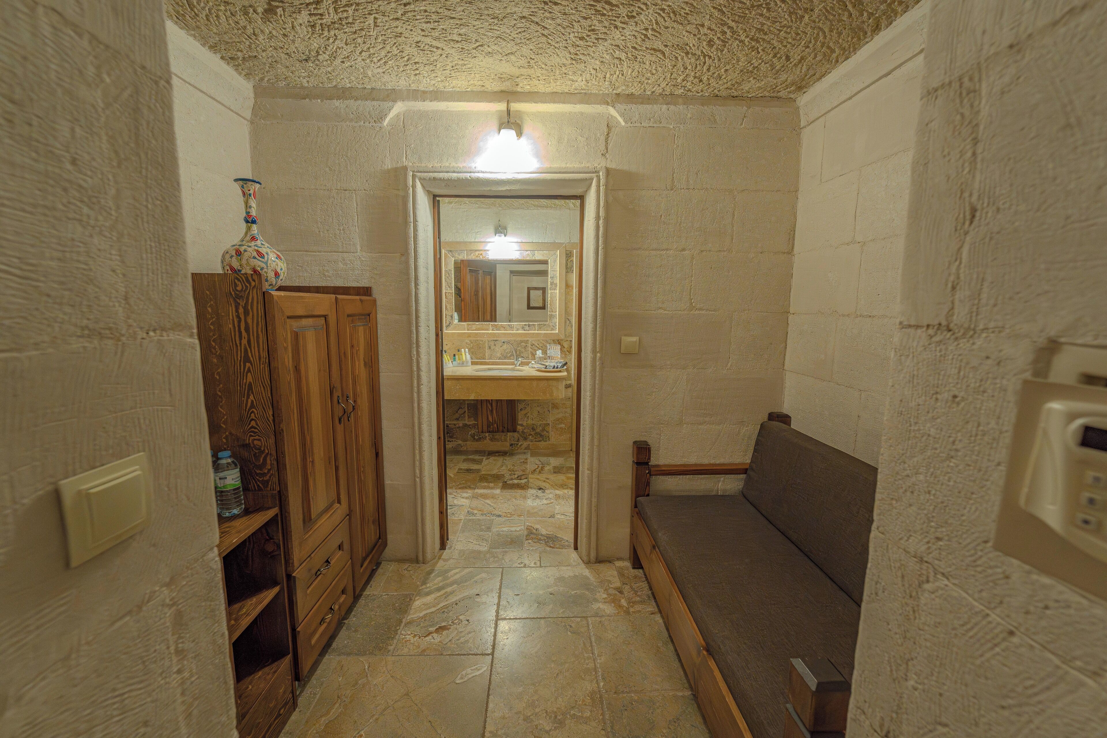 A narrow hallway with a wooden cabinet, a bench, and a mirror reflecting a bathroom with a sink and mirror.