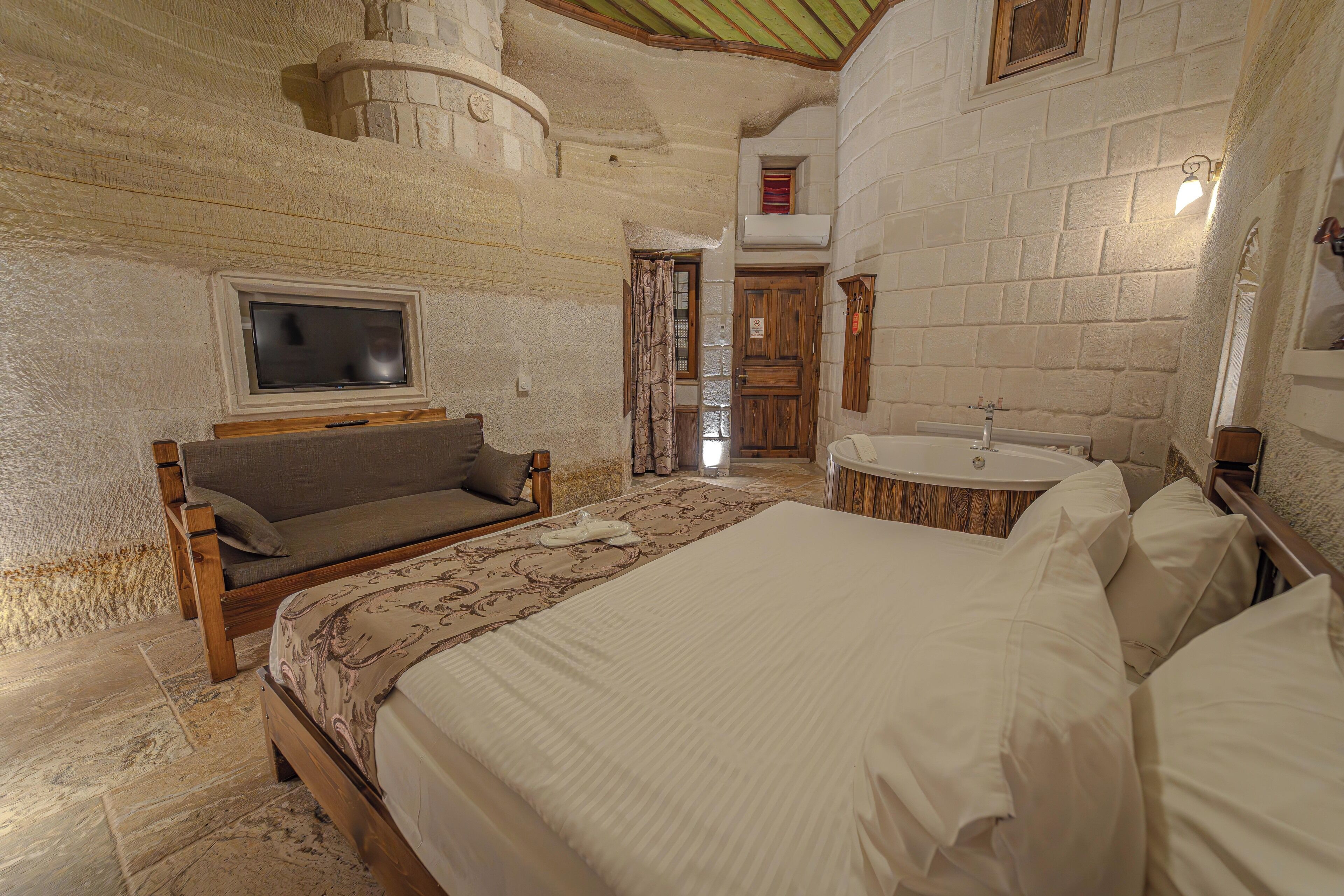 A bedroom with a bed, a television, a wooden bench, a bathtub, and a stone wall.