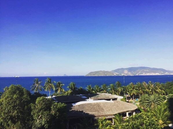A resort with thatched-roof huts, palm trees, and a view of the ocean and distant mountains. - Anh Minh Manchester (Nha Trang)