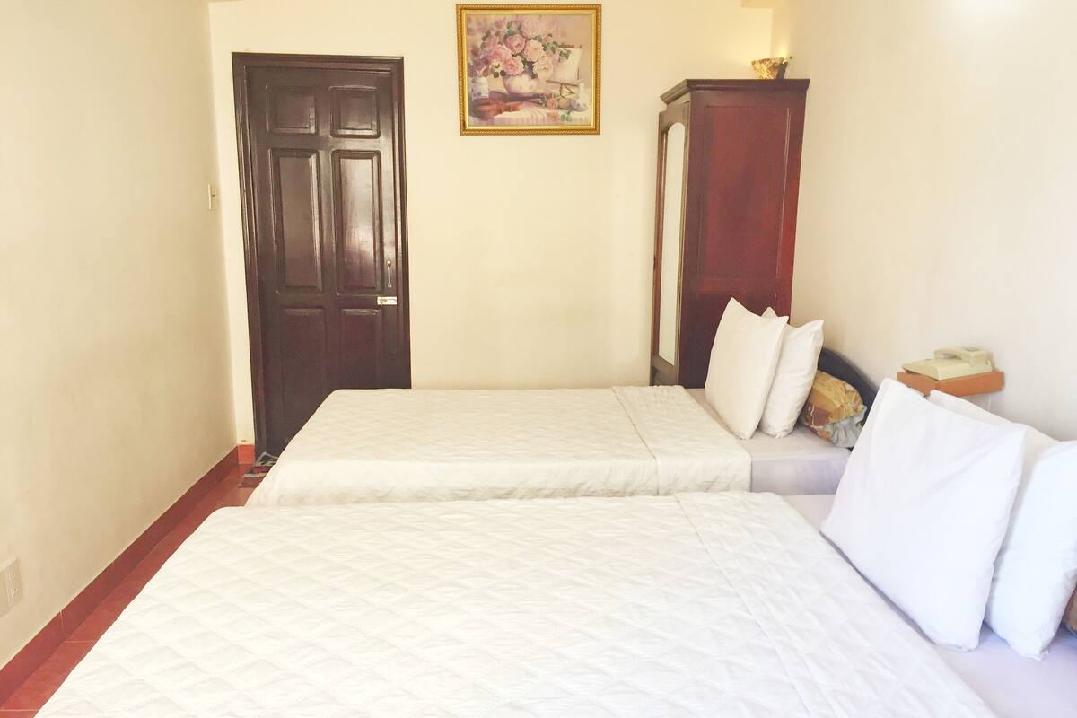 A hotel room with two beds, a wooden door, a painting on the wall, and a small table with a lamp.