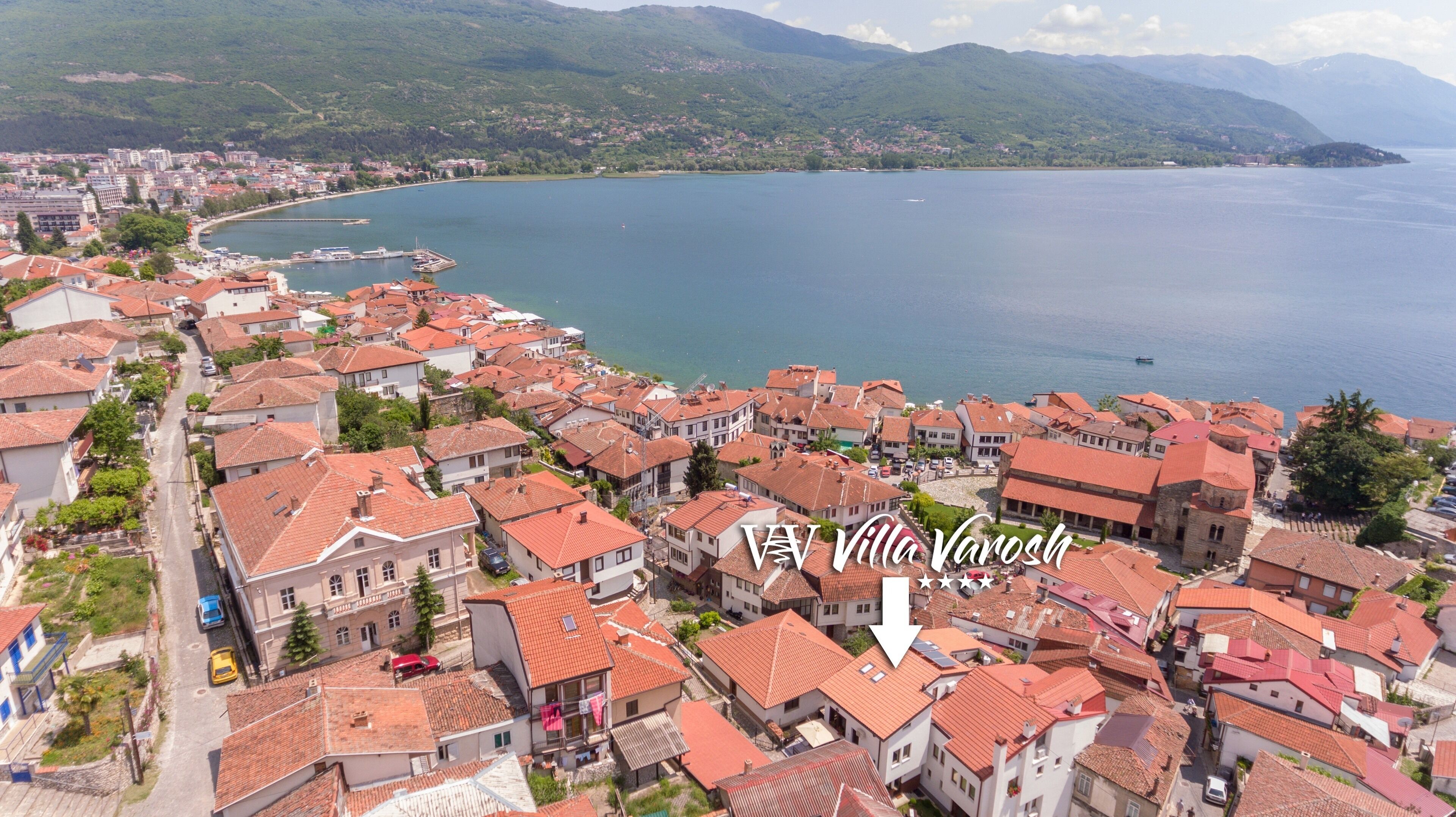 An aerial view of a coastal town with red-tiled roofs, a marina, and mountains in the background.
