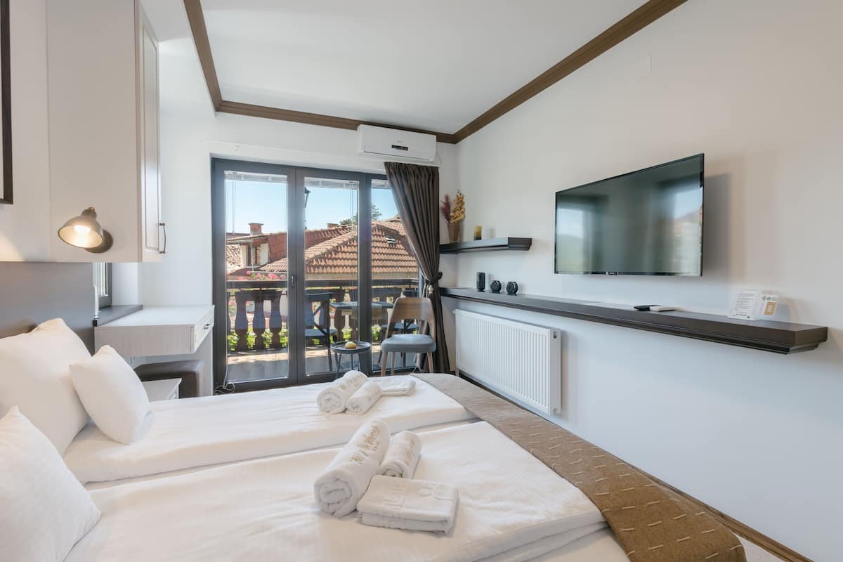 A hotel room with two beds, a TV, a desk, and a balcony with a view of buildings.