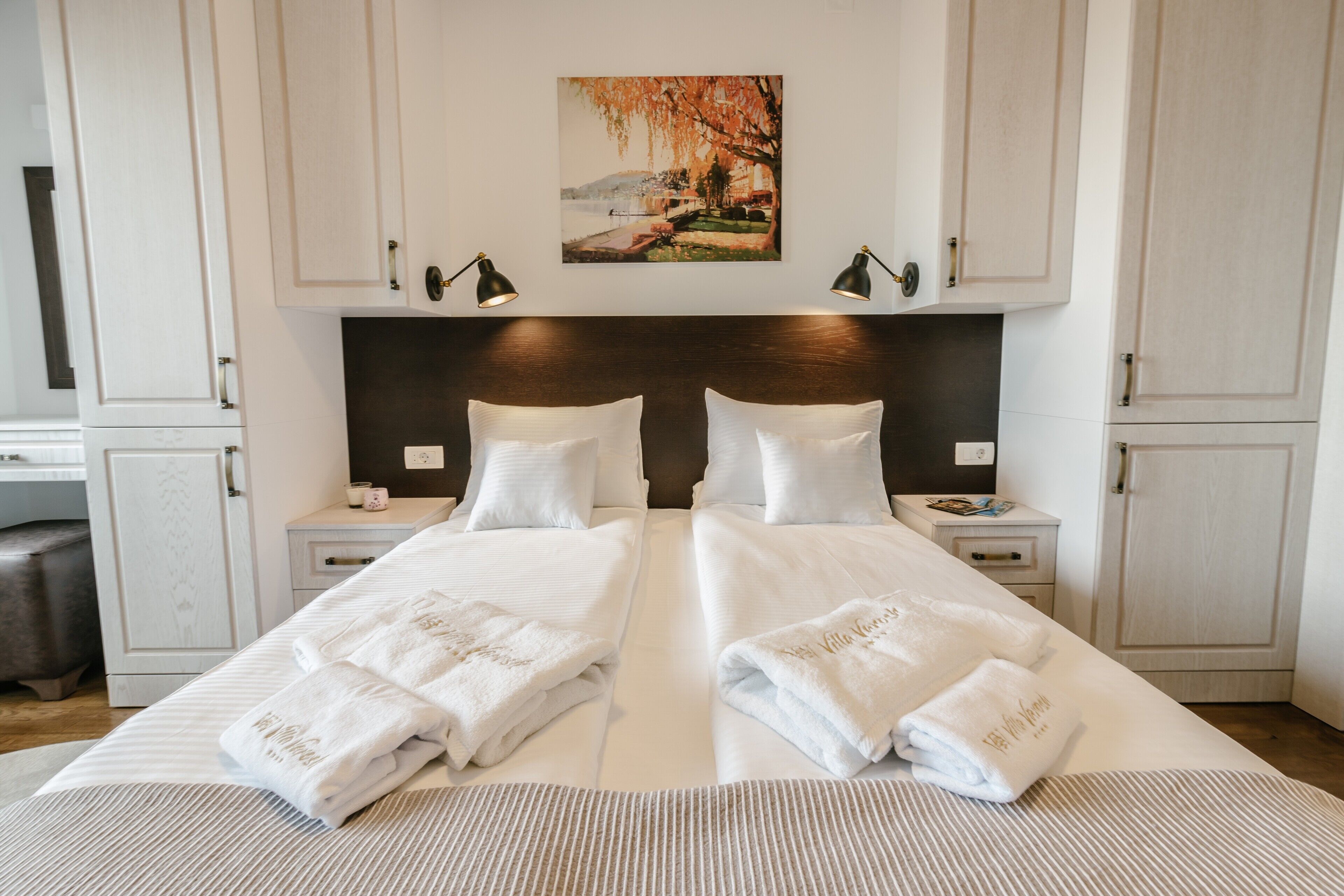 A double bed with white linens and towels, a painting above the headboard, and a dark wood headboard.