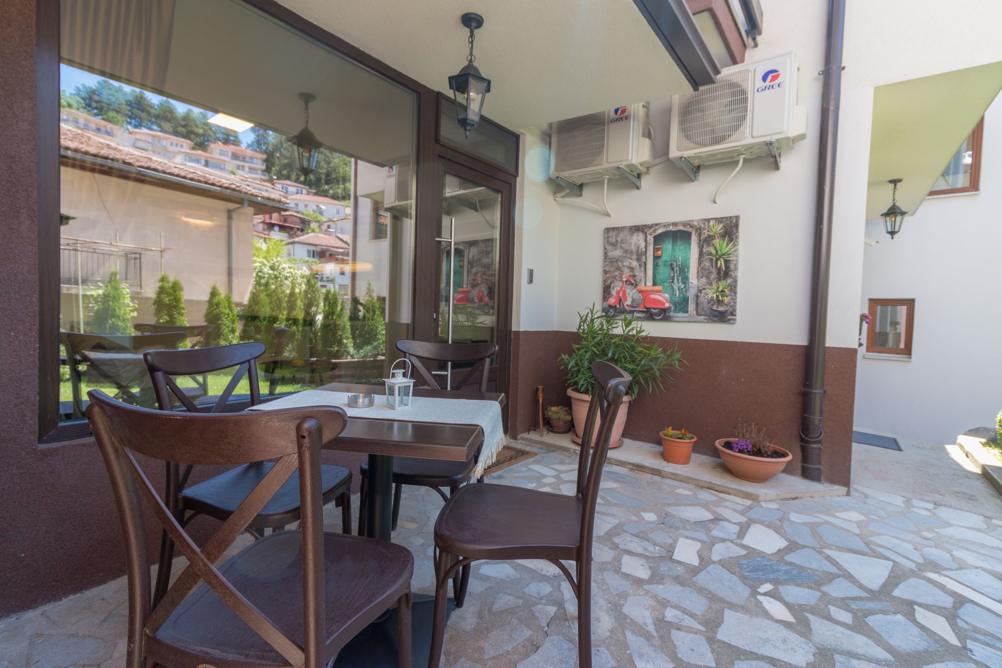 Un área de comedor al aire libre con suelo de piedra, sillas de madera y mesas. Se observan unidades de aire acondicionado en la pared, y macetas con plantas decoran el espacio de asiento.