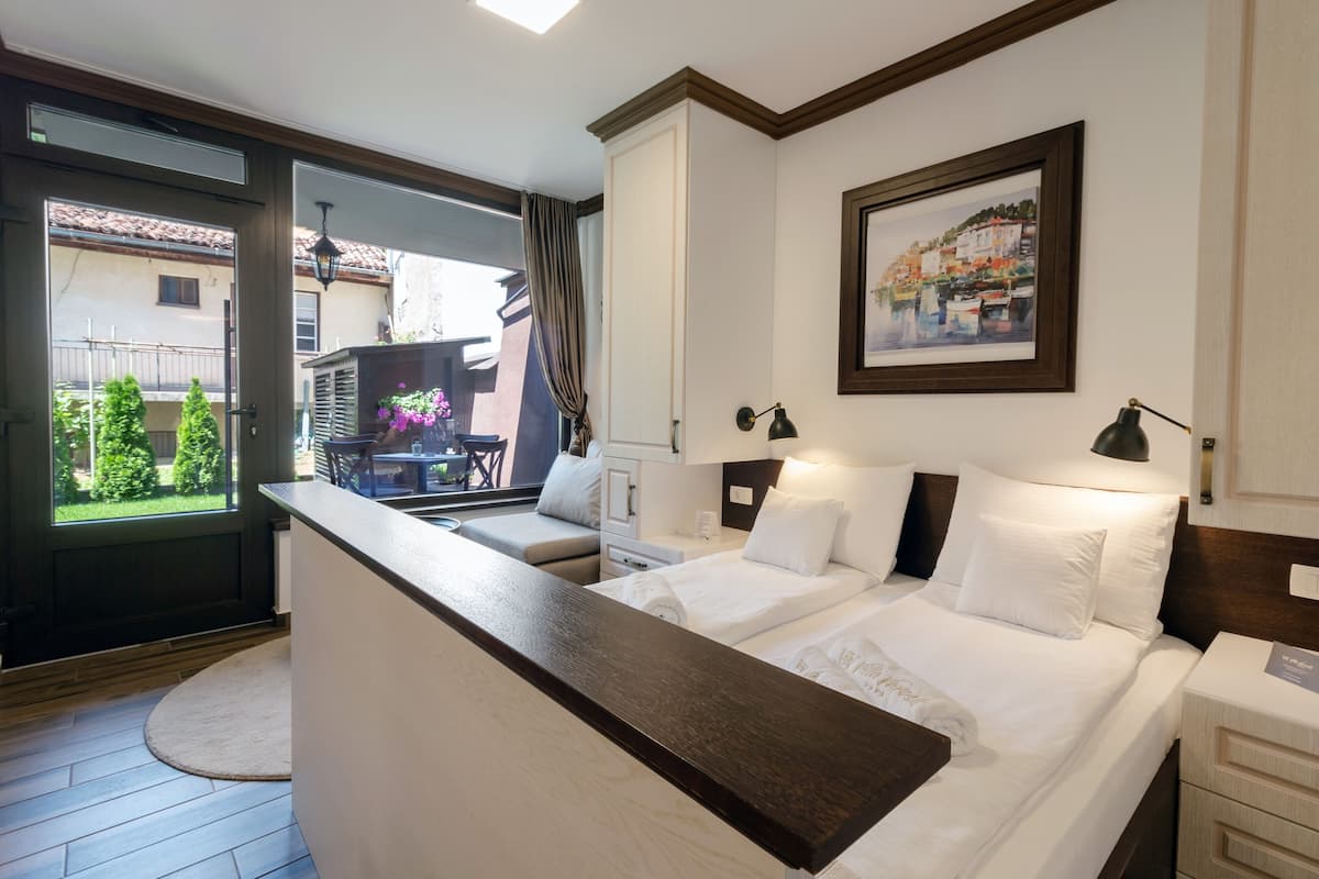A hotel room with a bed, bedside tables, a desk, a window with a view of greenery, and a painting on the wall.