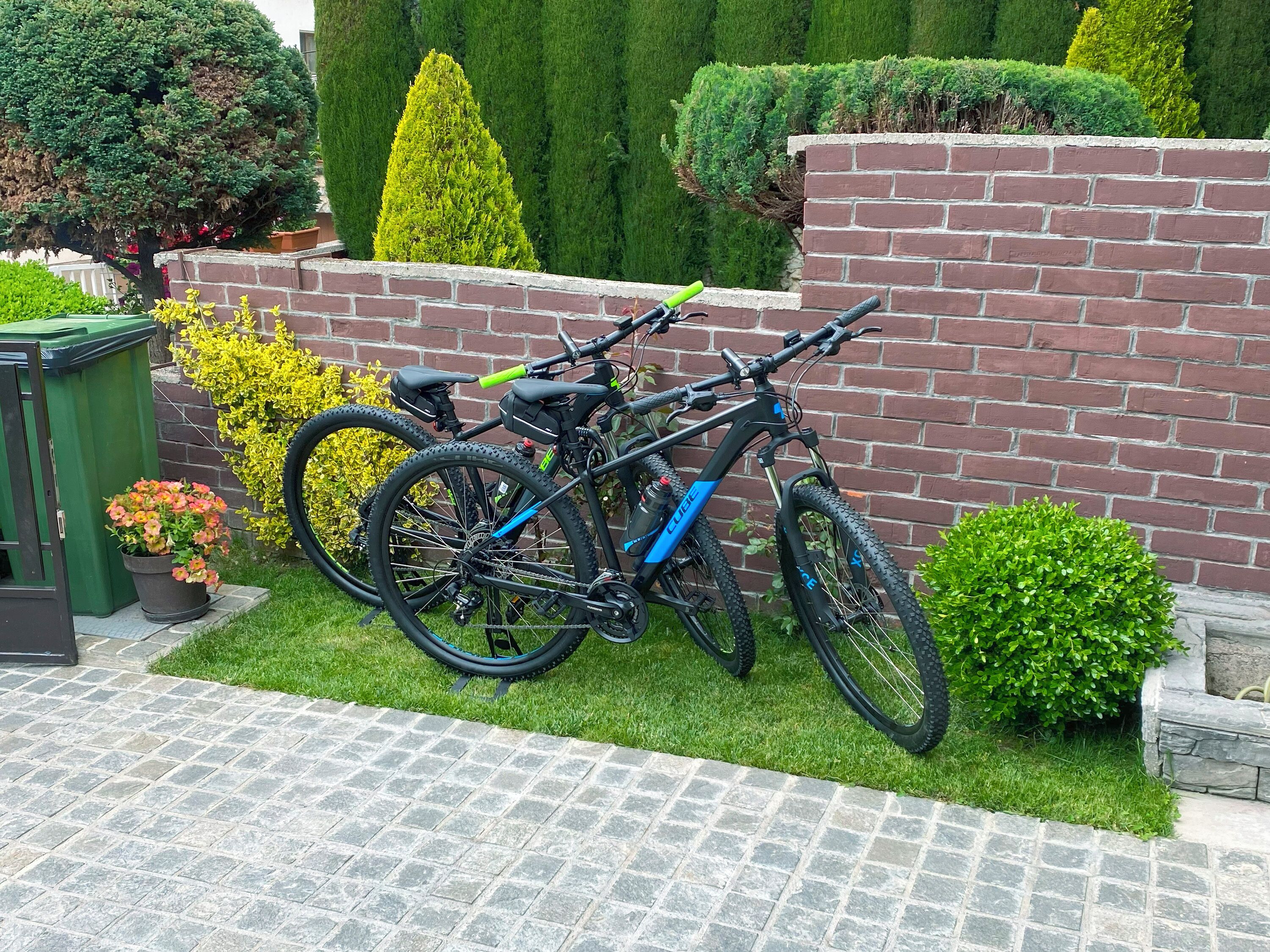 Twee fietsen staan geparkeerd tegen een bakstenen muur in een tuin met plantenpotten en een geplaveid pad.