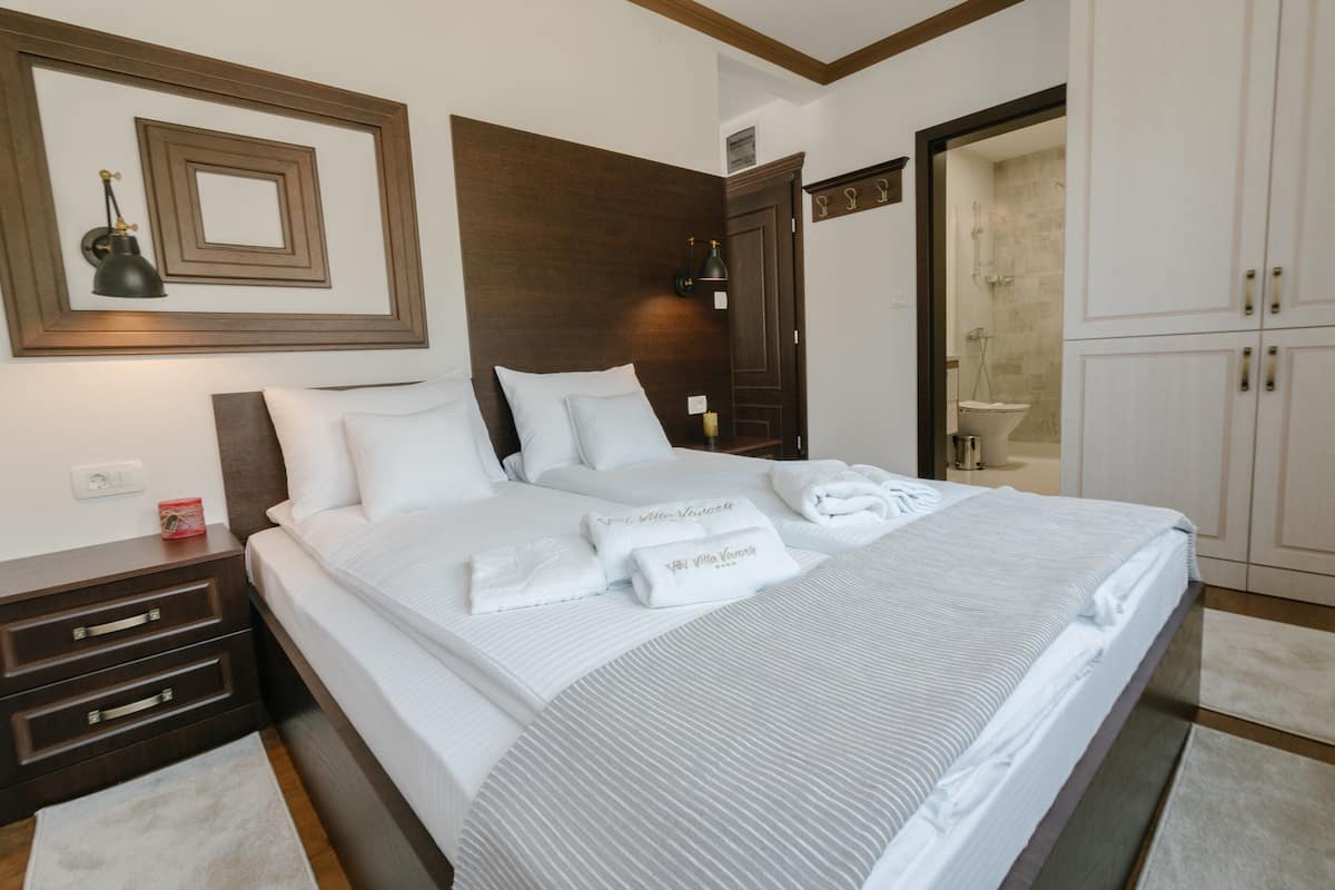 A neatly made bed with white linens and towels, a wooden headboard, a nightstand with drawers, and a wall-mounted lamp.