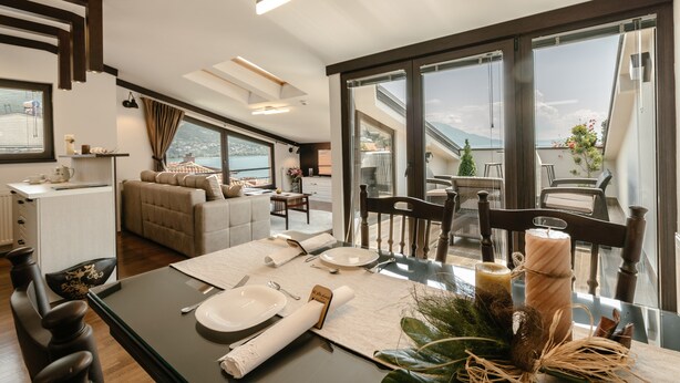 Un comedor moderno con una mesa de vidrio preparada para cuatro personas, un sofá al fondo y vistas al exterior a través de amplios ventanales.