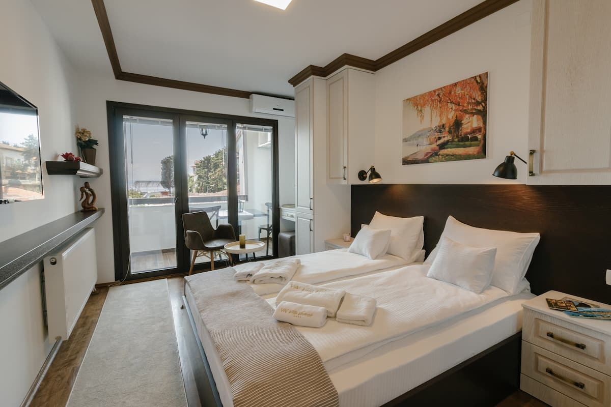 A hotel room with two beds, a large window, a balcony, and a painting on the wall.