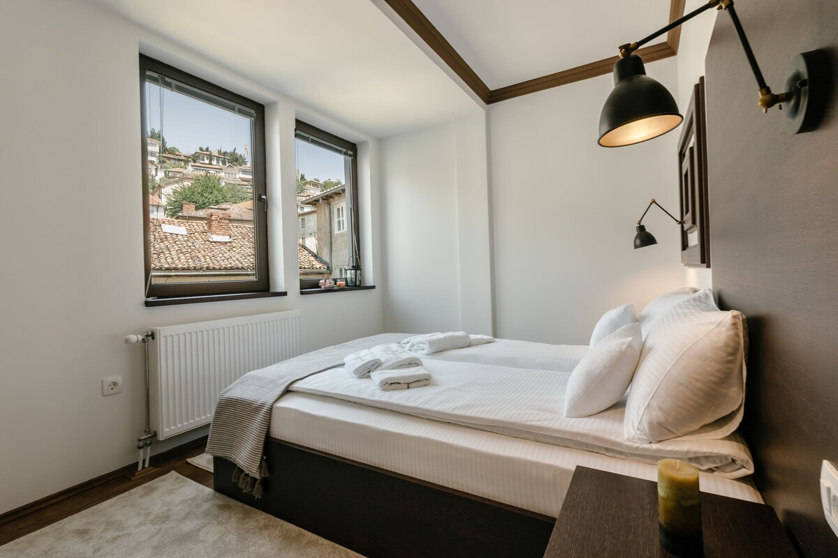 Un dormitorio con cama, dos ventanas, un radiador, una mesita de noche con vela y vistas a edificios.