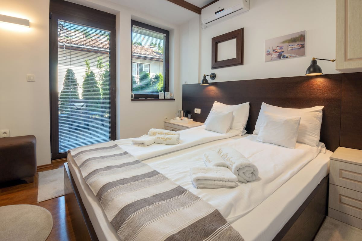 A hotel room with a large bed, two bedside tables, a nightstand, a chair, a window with blinds, and a wall-mounted air conditioner.