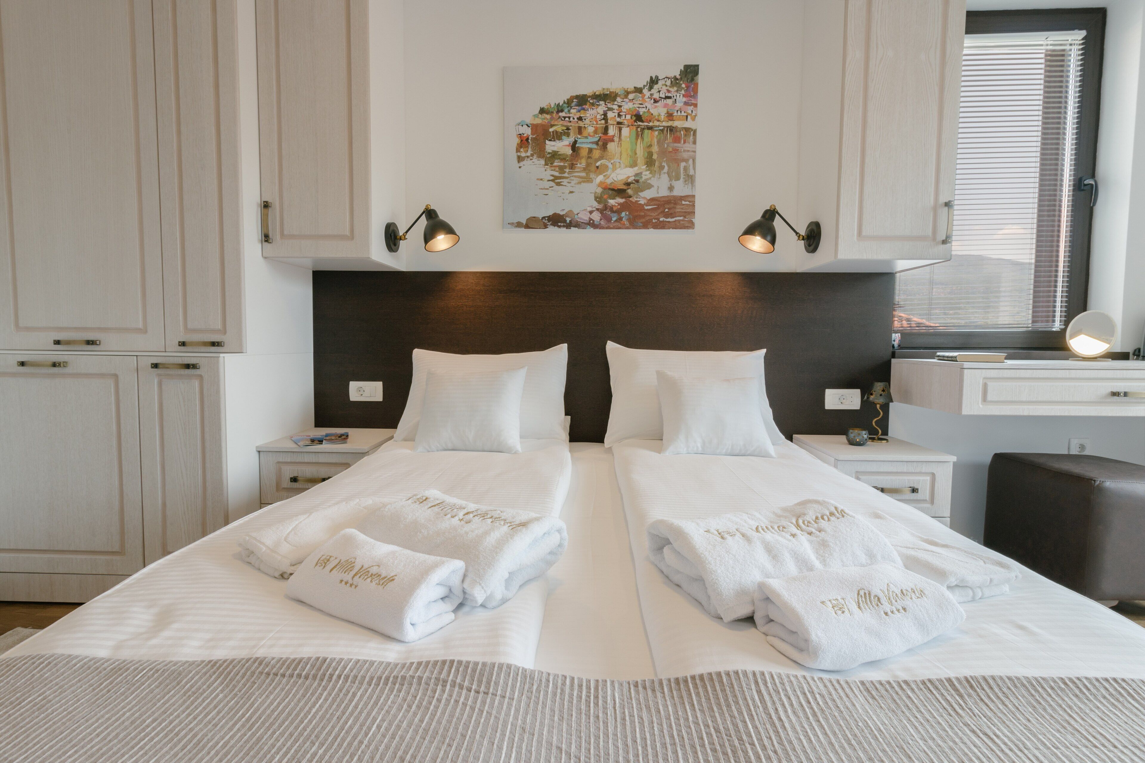 A hotel room with two beds, white linens, a painting of a coastal scene, and a desk with a lamp.