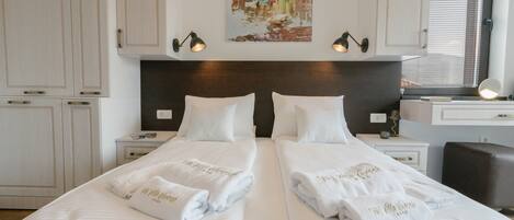 A hotel room with two beds, white linens, a painting of a coastal scene, and a desk with a lamp.