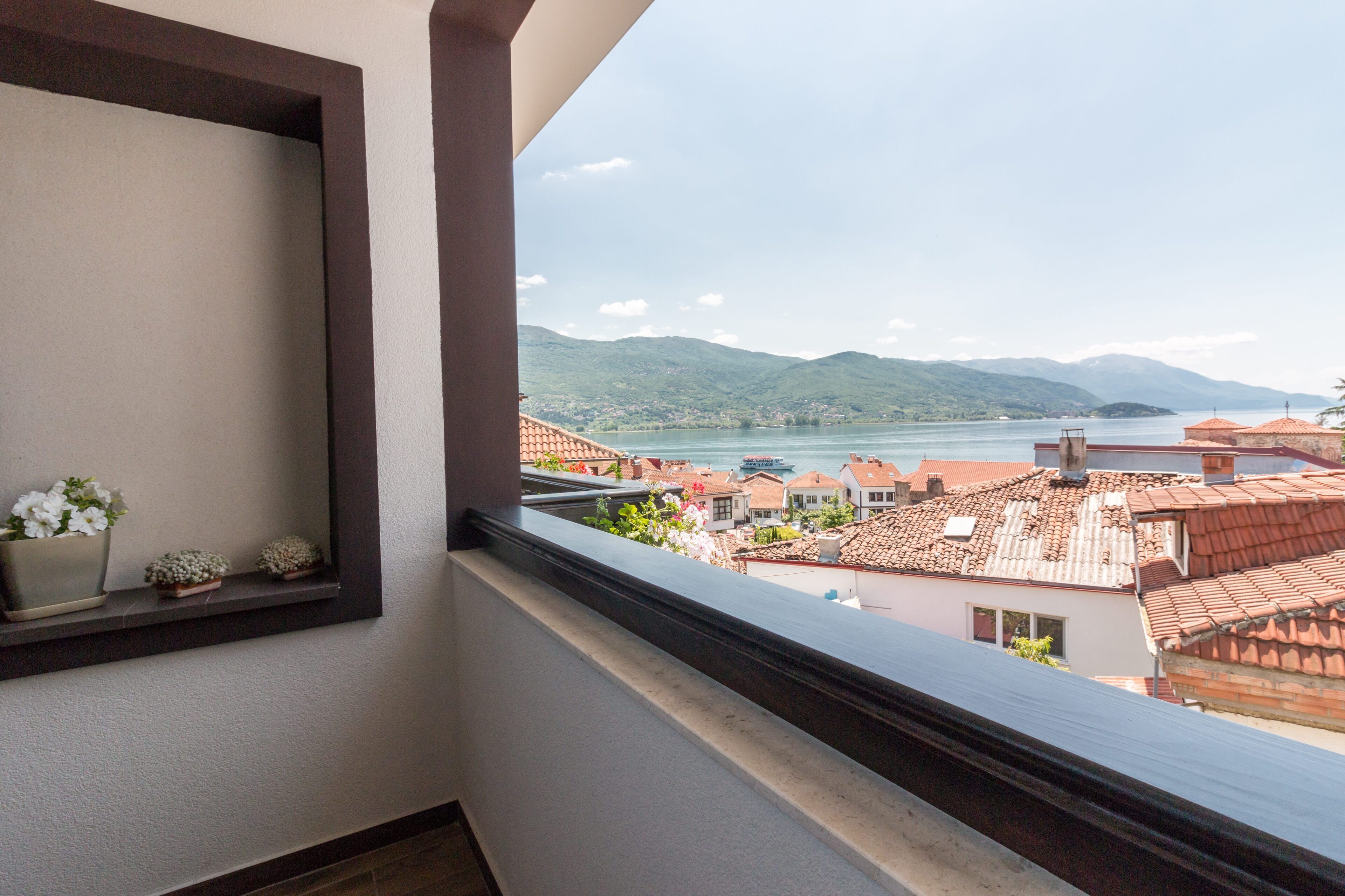 A balcony with a view of a lake and mountains, a shelf with flowers, and a tiled roof in the background.