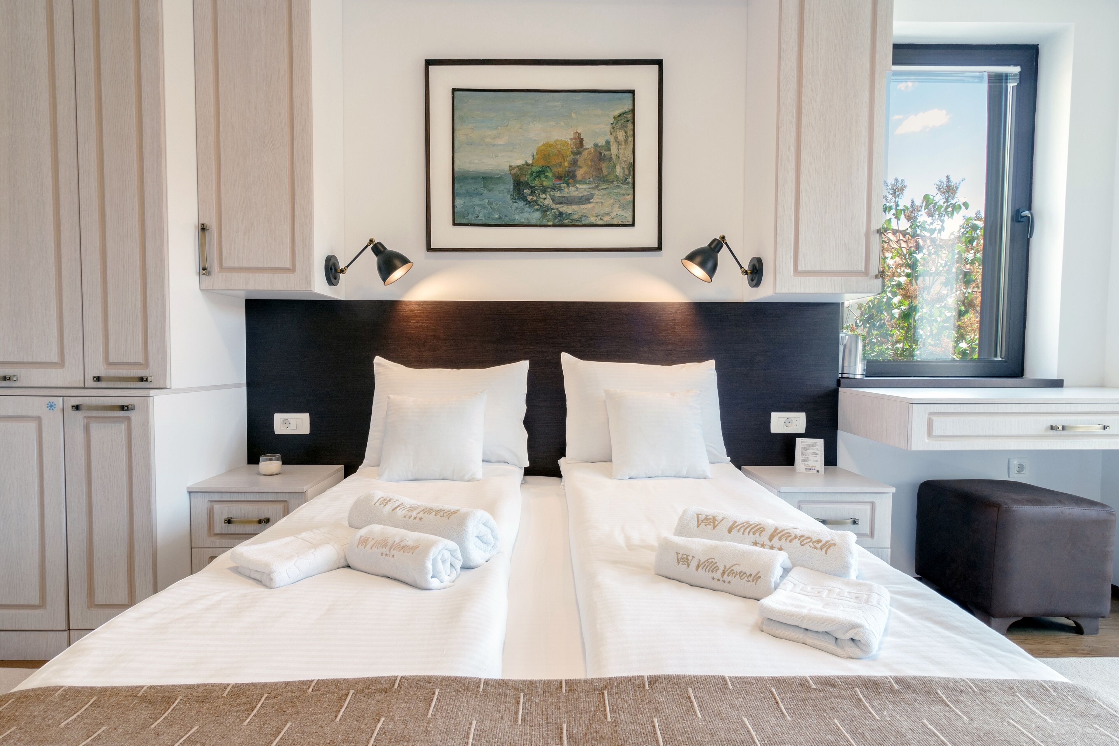 Una cama doble con ropa de cama y toallas blancas, un cuadro sobre la cama y una ventana con vistas a árboles.