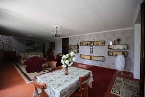 A dining area with a table set for four, surrounded by wooden chairs. A vase with flowers is on the table. The room has patterned wallpaper and a chandelier.