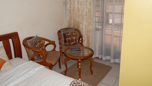 Une chambre à coucher avec un lit, deux chaises en bois, une petite table ronde et une fenêtre ornée de rideaux légers.