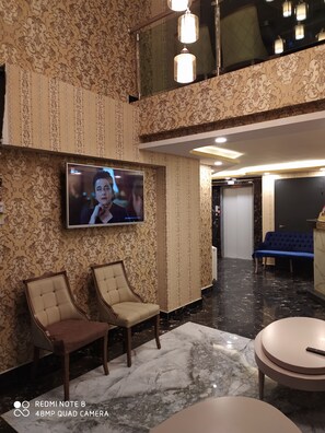 Un vestíbulo con papel tapiz estampado, un televisor montado en la pared, dos sillones tapizados, un suelo de mármol y un mostrador de recepción.