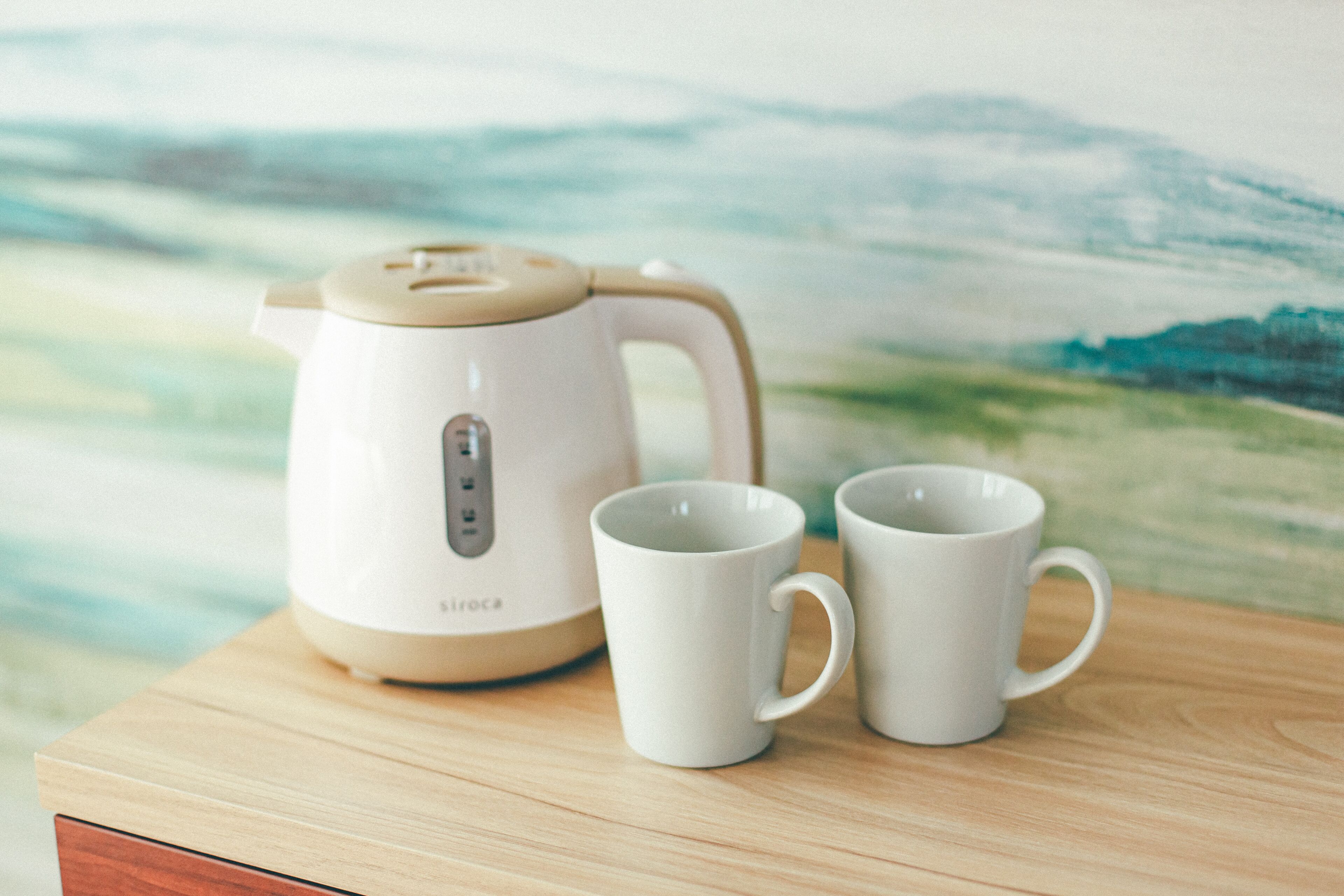 A white electric kettle and two white mugs on a wooden table with a blurred landscape painting in the background.