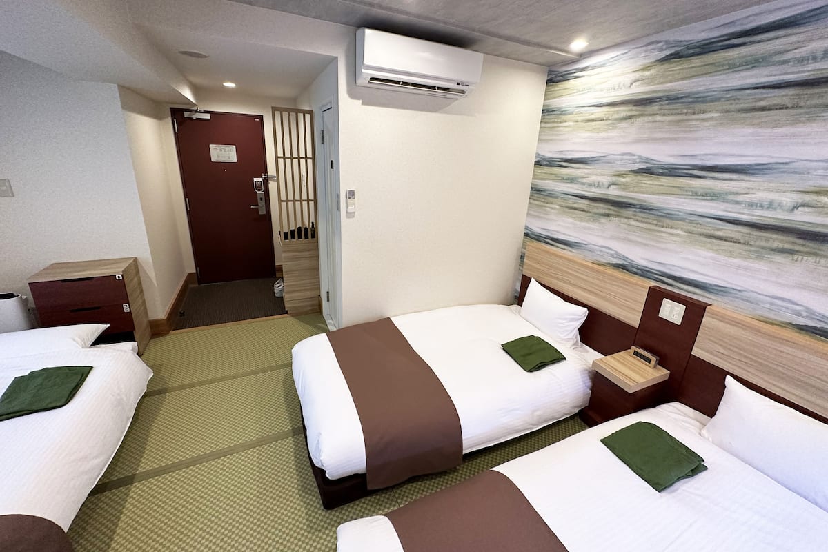 Une chambre d’hôtel avec deux lits, une tête de lit en bois, une moquette à motifs verts, un appareil de climatisation et un tableau au mur.