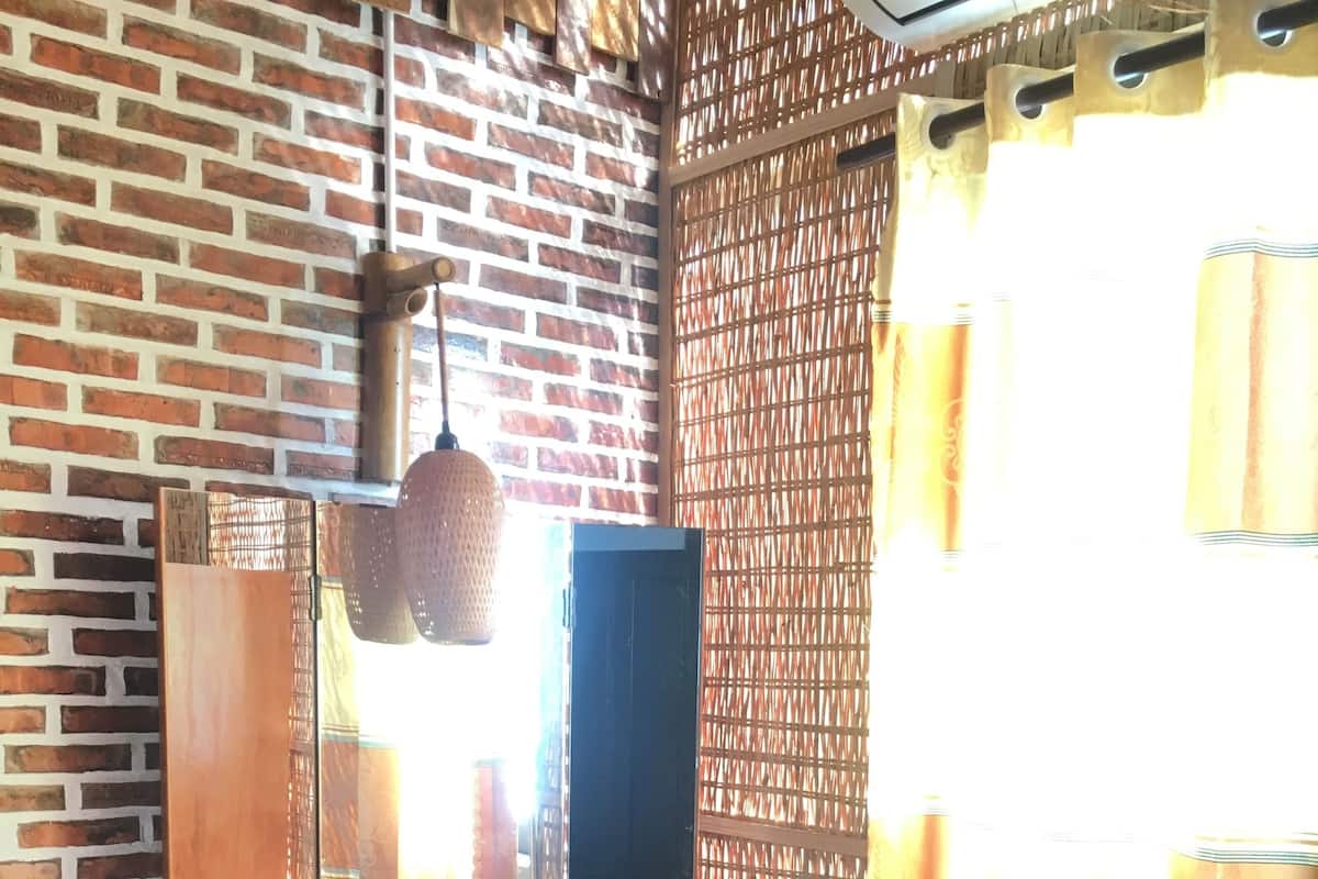 A room with a brick wall, a wooden desk with a mirror, a chair, and a window with patterned curtains.