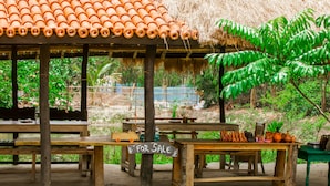 A rustic outdoor seating area with wooden tables and benches, a thatched roof, and a 'For Sale' sign. - Backpackers Eco Village (Gosaba)