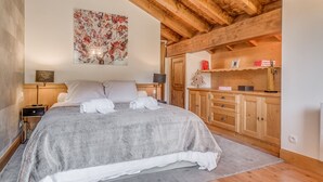 Une chambre à coucher avec un grand lit, des poutres apparentes en bois au plafond, une table de chevet, une commode, un tableau accroché au mur et une armoire en bois dotée d’un miroir.