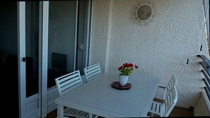 Un balcón con una mesa y sillas blancas, una planta en maceta con flores rojas y un adorno en la pared.
