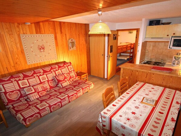 Een gezellige kamer met een houten vloer, een bank met een hoesje met een hartmotief, een eettafel met een feestelijk tafelkleed en een keukenhoek met een fornuis en kastjes.