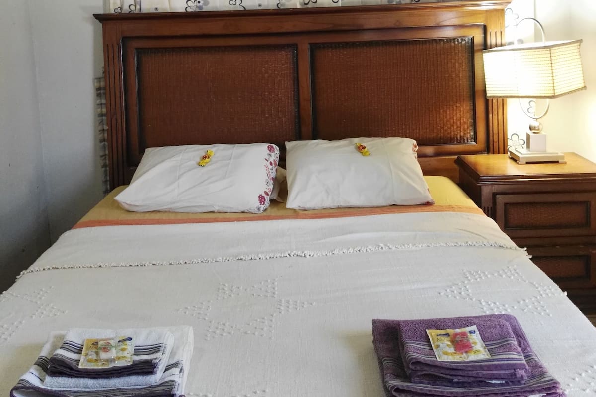 A hotel room with a bed, pillows, a nightstand, a lamp, and a folded towel on the bed.